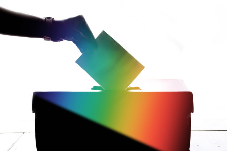 a hand placing a ballot into a rainbow colored ballot box